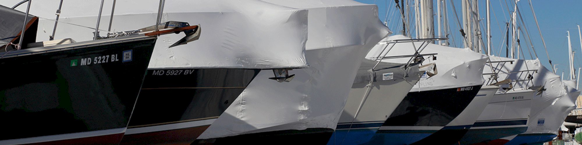 Several boats covered in protective tarps are lined up on a snowy surface in a marina, with masts extending upwards.