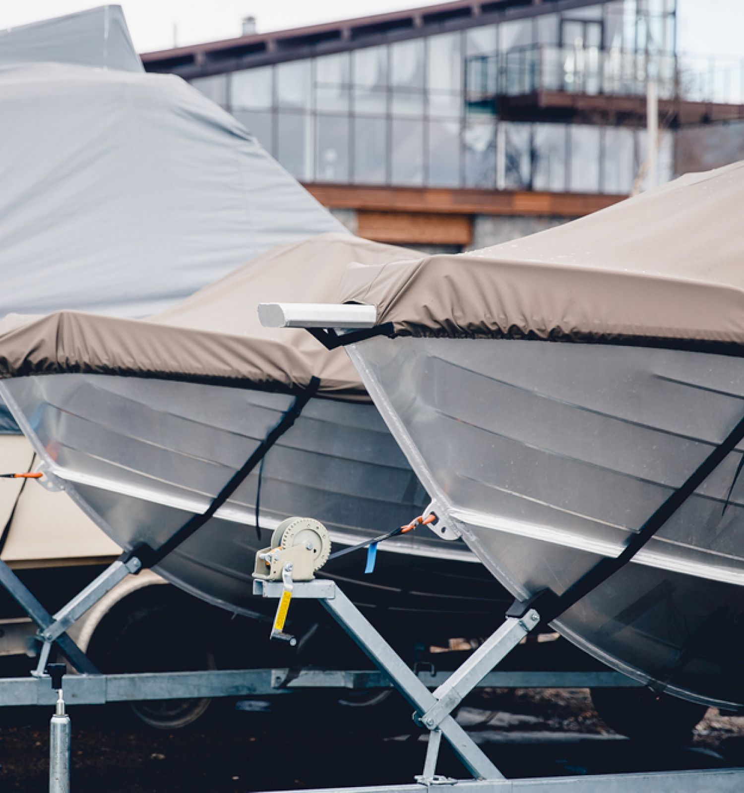 The image shows two covered boats on trailers, parked outdoors with a building in the background.