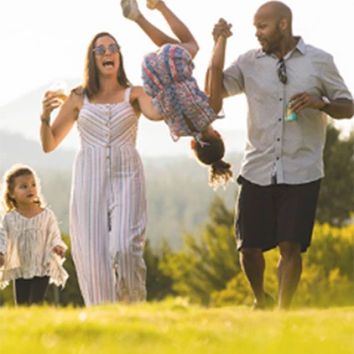 A family of four enjoys the outdoors, with the parents lifting one child playfully in the air while the other child walks beside them.