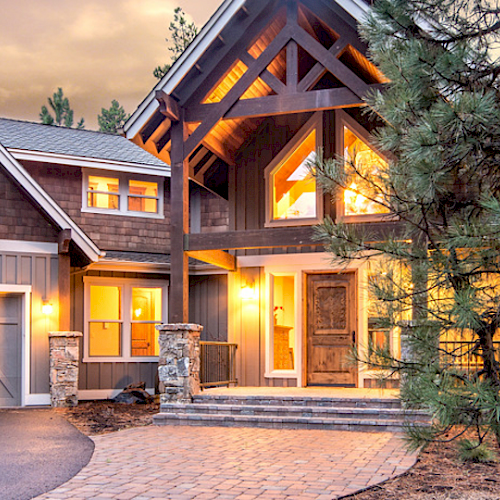 A large, illuminated modern house with a wooded exterior and a driveway, surrounded by trees during twilight or sunset.