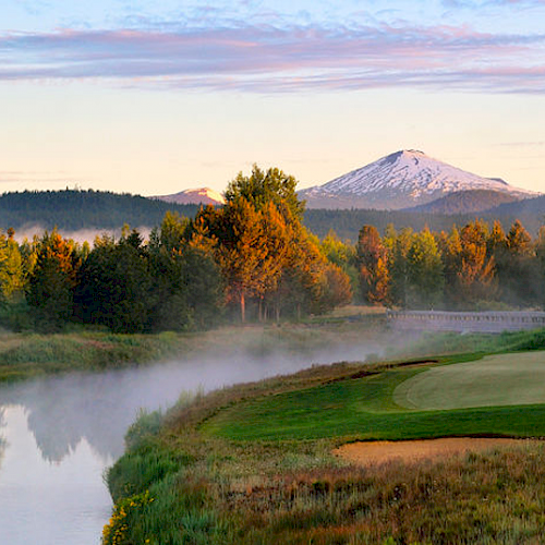The image shows a scenic view of a golf course next to a river, with a flag on the green, trees, and a snow-capped mountain in the background.