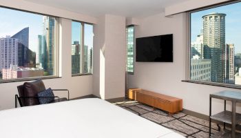 A modern bedroom with large windows, a bed, a chair, a TV on the wall, and a cityscape view of tall buildings outside.