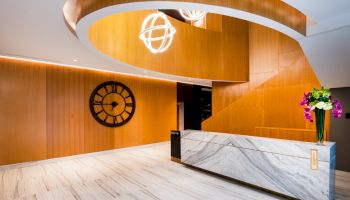 The image shows a modern lobby with wooden walls, a large clock, a spiral staircase, a marble reception desk, and a flower arrangement.