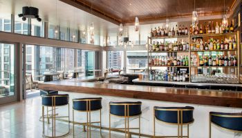 The image shows a modern bar with a wooden counter, blue and gold bar stools, and a well-stocked shelf of bottles.