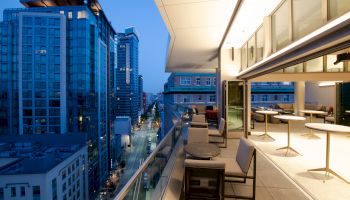 A modern balcony overlooking a city street with tall buildings, featuring outdoor seating and tables under a roof. The city is lit up at dusk.