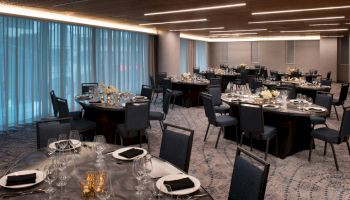 The image shows an elegantly set banquet hall with round tables and chairs, dressed with plates, glassware, black napkins, and floral centerpieces.