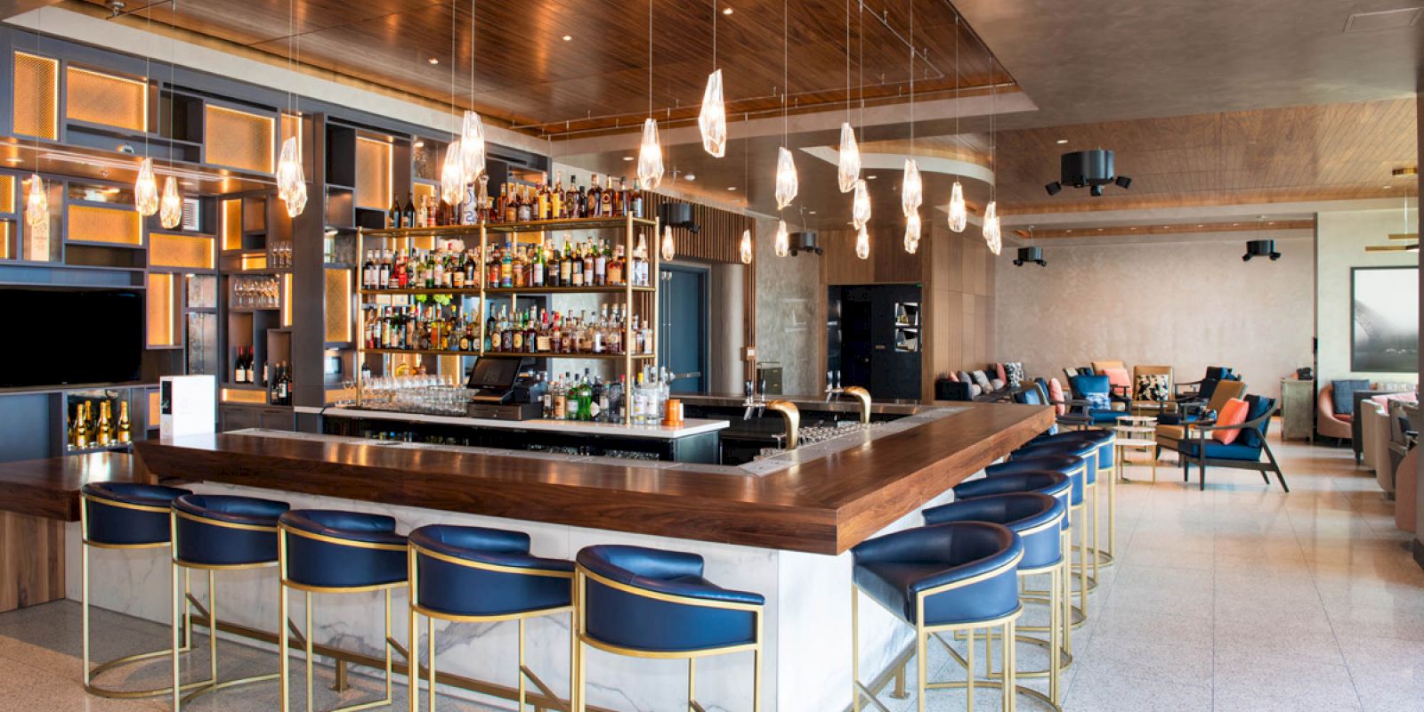 A modern bar with blue stools, a wooden countertop, hanging lights, and a well-stocked liquor shelf. There's seating in the background.