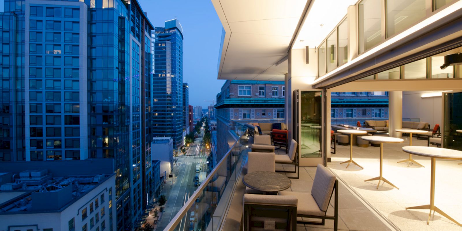 The image shows a modern rooftop patio at dusk, with city skyscrapers in the background and outdoor seating arrangements on the right.