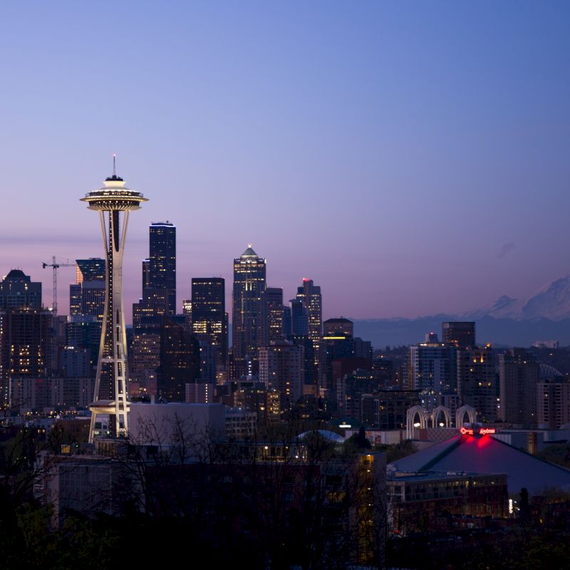 A twilight view of a city skyline featuring the Space Needle prominently, with Mount Rainier visible in the background against a clear sky.