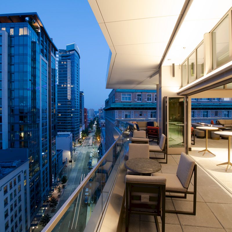 A modern balcony with tables and chairs overlooks a city street lined with tall buildings at twilight, featuring a sleek and open design.