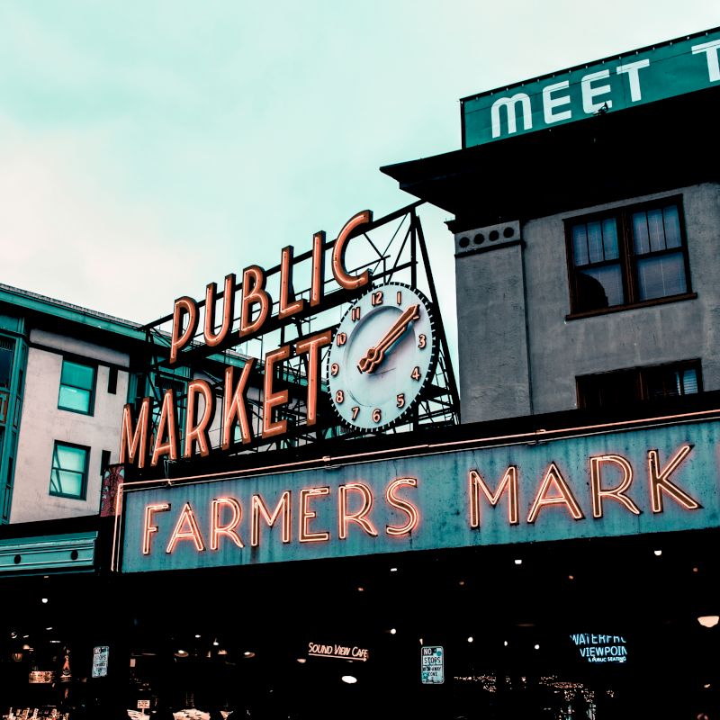 This image shows signage for a "Public Market" and "Farmers Market" with a clock, and surrounding buildings in an urban area.