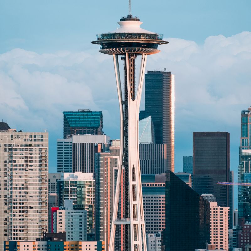 The image features the Space Needle in Seattle, with various modern buildings and skyscrapers in the background under a partly cloudy sky.