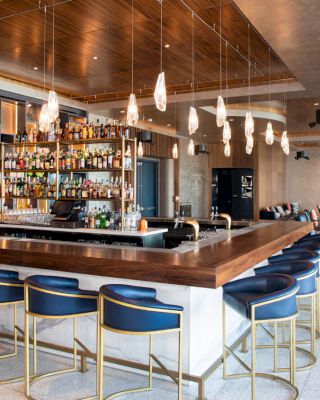 The image shows a stylish bar interior with a central counter, blue bar stools, and pendant lights. Bottles of alcohol are displayed behind the bar.