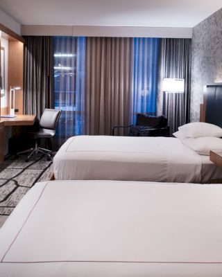 A modern hotel room with two double beds, a flat-screen TV, a desk with a chair, and contemporary decor, including patterned walls and carpeting.