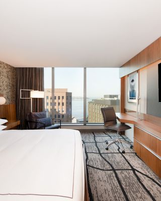 A modern hotel room with a large bed, wall-mounted TV, desk, and a view of the city through floor-to-ceiling windows.