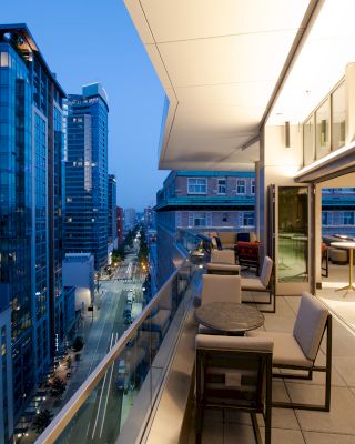 A rooftop balcony overlooks a cityscape with tall buildings at dusk. Tables and chairs are arranged on the balcony, facing large windows.