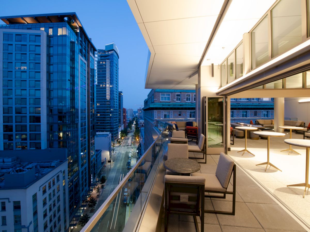 The image shows a modern cityscape view from a terrace at dusk with seating arrangements and buildings lining the street, under a clear sky.