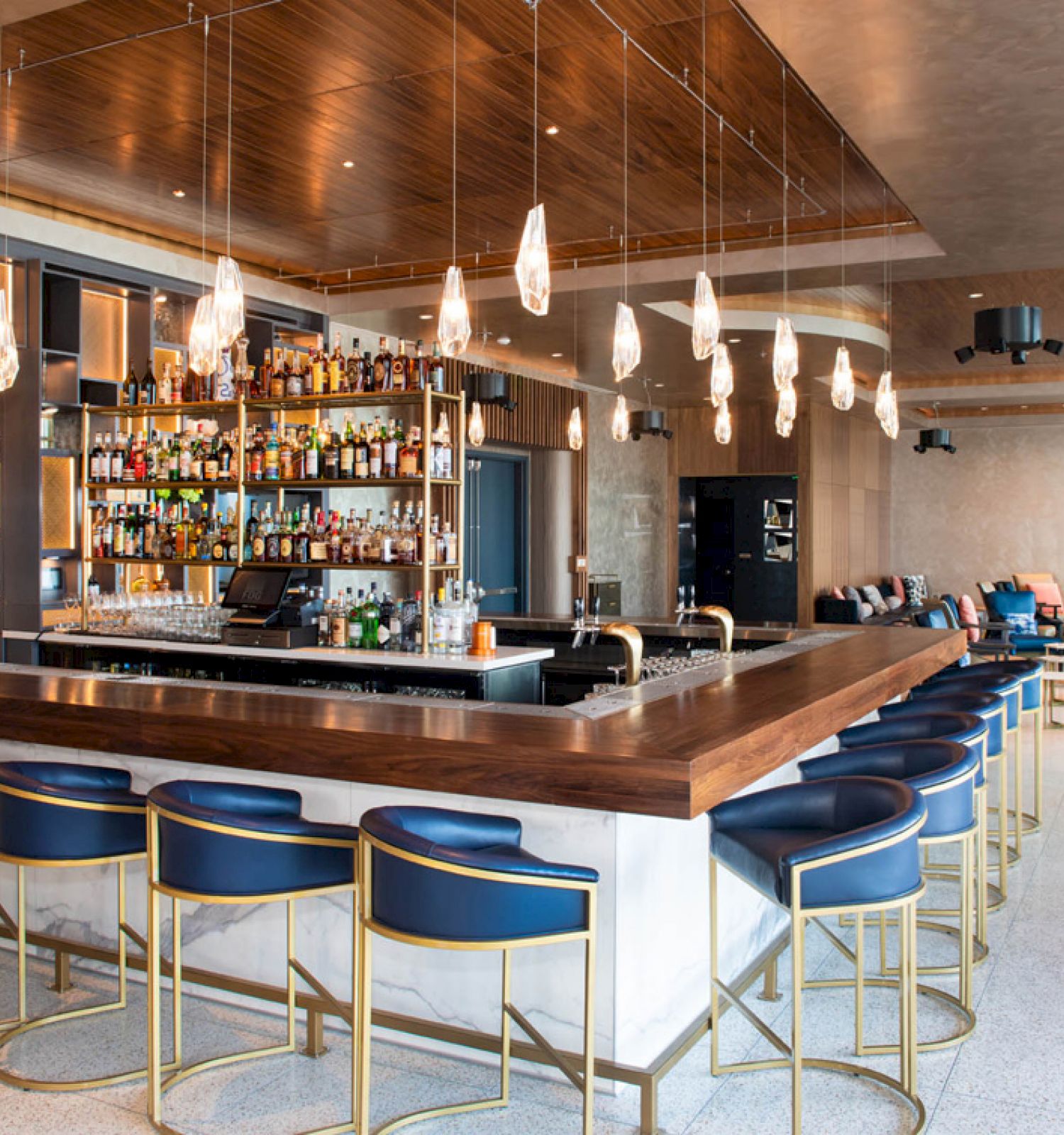 A modern bar with a stocked shelf, stylish pendant lights, and blue bar stools arranged around a wooden counter inside a chic lounge area.
