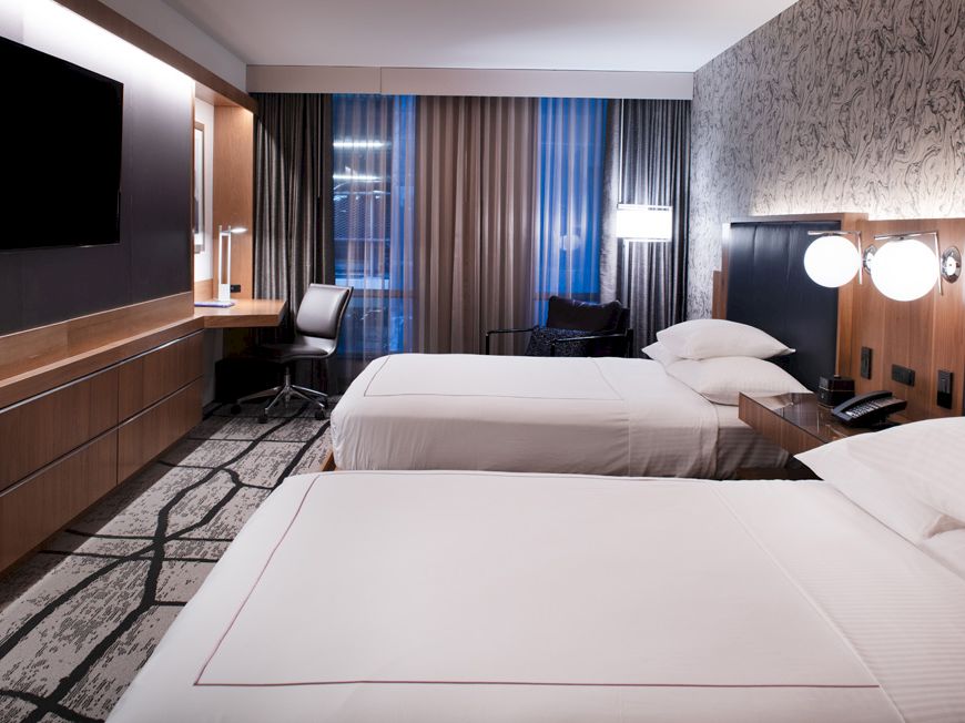 A modern hotel room with two neatly made beds, a wall-mounted TV, a desk with a chair, and stylish decor featuring wood and patterned wallpaper.