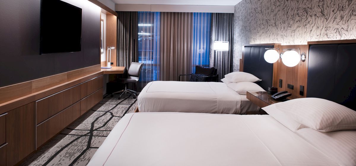 A modern hotel room with two neatly made beds, a wall-mounted TV, a desk with a chair, and stylish decor featuring wood and patterned wallpaper.