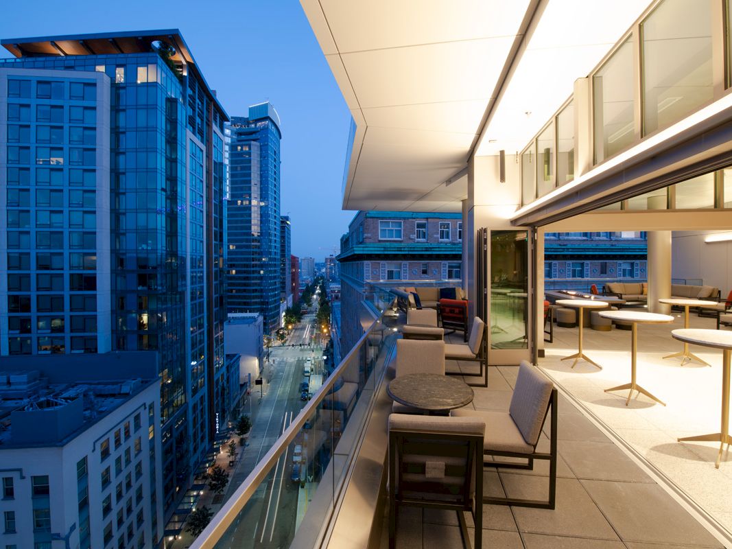 The image shows a modern balcony with seating and tables, overlooking a city skyline with tall buildings during the evening.