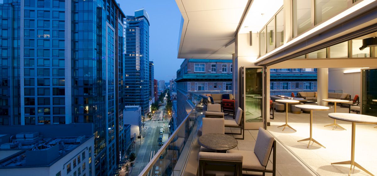 The image shows a modern balcony with seating and tables, overlooking a city skyline with tall buildings during the evening.