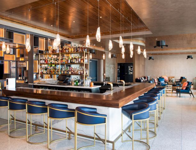 A modern, stylish bar with blue chairs and hanging lights, featuring a well-stocked drinks shelf and a cozy seating area in the background.