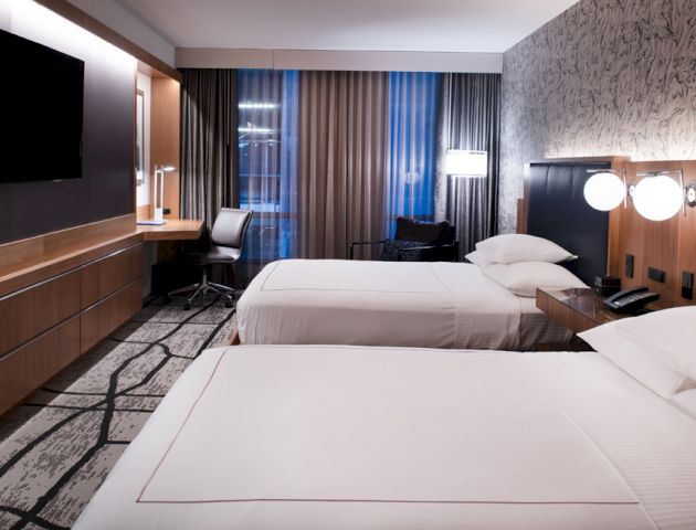A modern hotel room with two beds, a wall-mounted TV, a desk with a chair, contemporary lighting, and large windows with curtains.