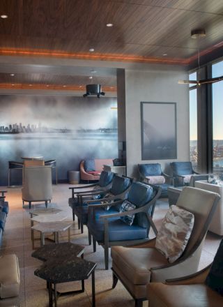 The image shows a modern lounge area with several comfortable chairs, tables, and large windows offering a scenic water view at dusk.