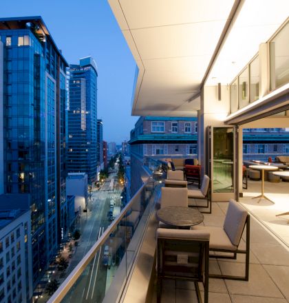 A modern balcony overlooks a city street lined with tall buildings at twilight, featuring tables, chairs, and an open indoor seating area.