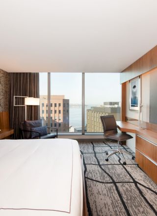 A modern hotel room with a large bed, wall-mounted TV, desk, chair, and a window showcasing a cityscape view.