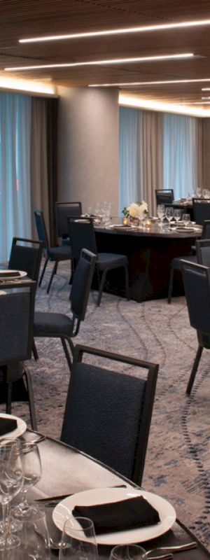 A banquet hall is set up for an event with round tables, black chairs, napkins, glassware, and floral centerpieces.