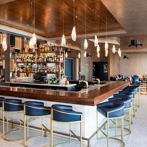 A modern bar with a stylish wooden counter, blue and gold bar stools, hanging lights, and a curved seating area with couches in the background.