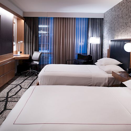 The image shows a modern hotel room with two neatly made beds, a wall-mounted TV, a work desk, and ambient lighting.