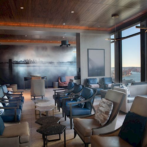 The image shows a modern lounge area with comfortable seating, large windows with a waterfront view, and a sleek design with a cityscape mural.