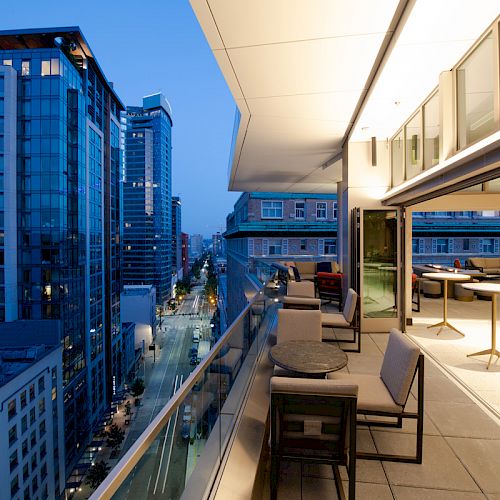 A modern cityscape view at dusk with tall buildings, seen from a spacious balcony or terrace with seating and tables positioned for relaxation.