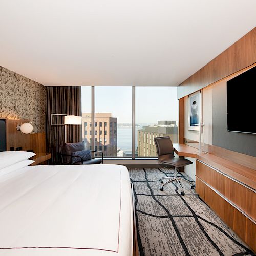 A modern hotel room with a large bed, wall-mounted TV, desk, chair, and a window revealing a cityscape view.