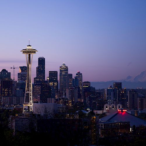 A city skyline at dusk featuring the Space Needle and downtown buildings, with a mountain in the background.
