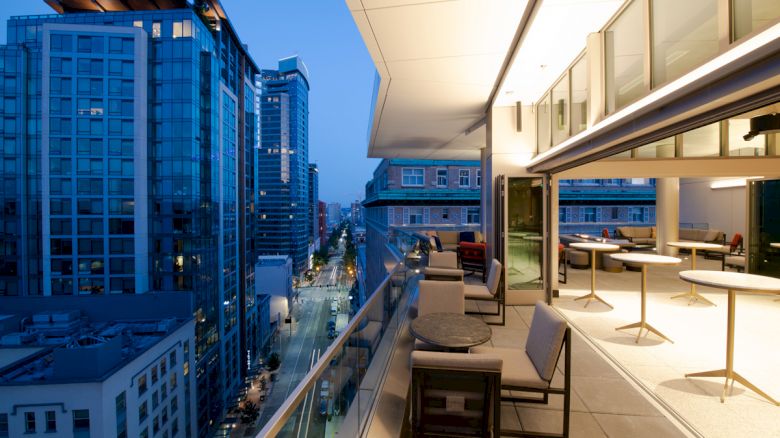 A modern balcony at dusk with tables and chairs, overlooking a cityscape of tall buildings and a street lined with streetlights and cars.