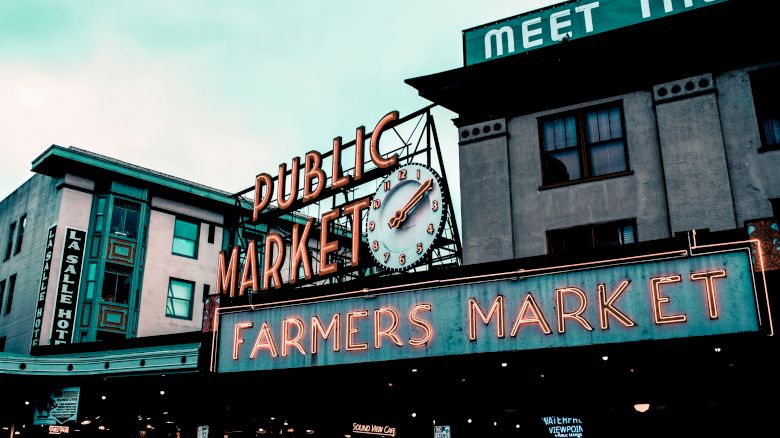 Neon signs reading "Public Market" and "Farmers Market" are displayed on a building against an overcast sky, with additional signage partially visible.