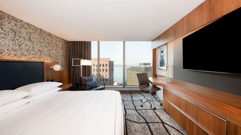Modern hotel room with a large bed, desk, chair, wall-mounted TV, and a view of buildings and water through floor-to-ceiling windows.