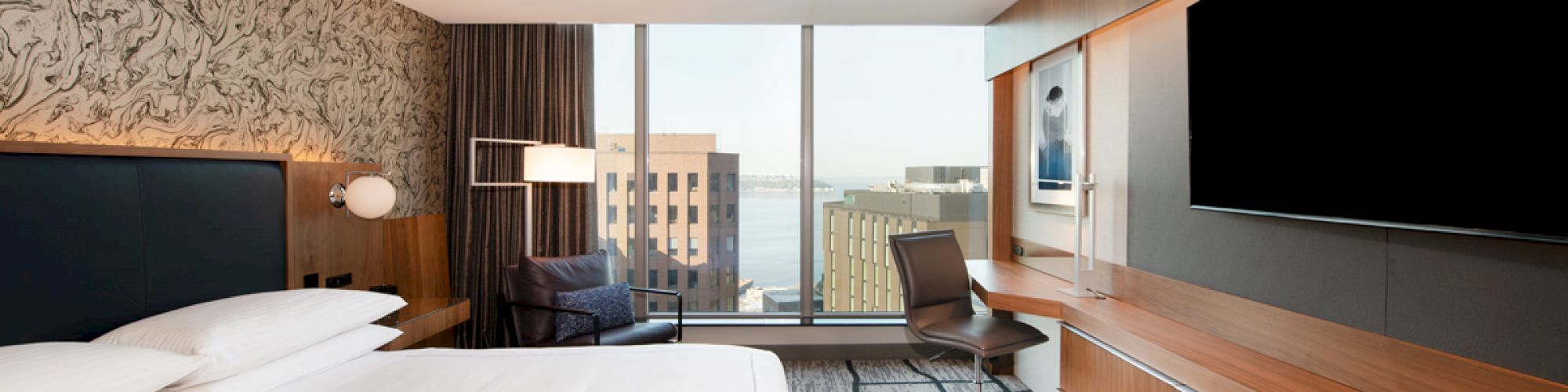 A modern hotel room with a king-sized bed, a desk with a chair, a large flat-screen TV, and a window with a city view in the background.