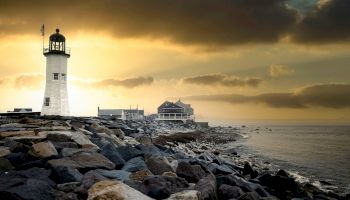 A lighthouse stands on a rocky coastline with buildings in the background under a dramatic, cloudy sky during sunset near a calm sea.