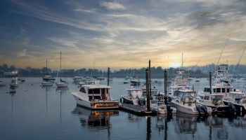 The image shows a serene harbor at dusk with several boats docked at the piers, calm water, and a picturesque sky.