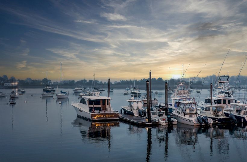 The image shows a serene harbor at dusk with several boats docked at the piers, calm water, and a picturesque sky.