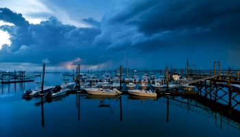 A serene marina at dusk, with boats docked and dramatic, cloudy skies reflecting on the calm water.