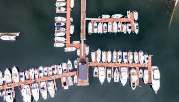 The image shows an aerial view of a marina with numerous boats docked at wooden piers extending into the water, surrounded by calm waters.