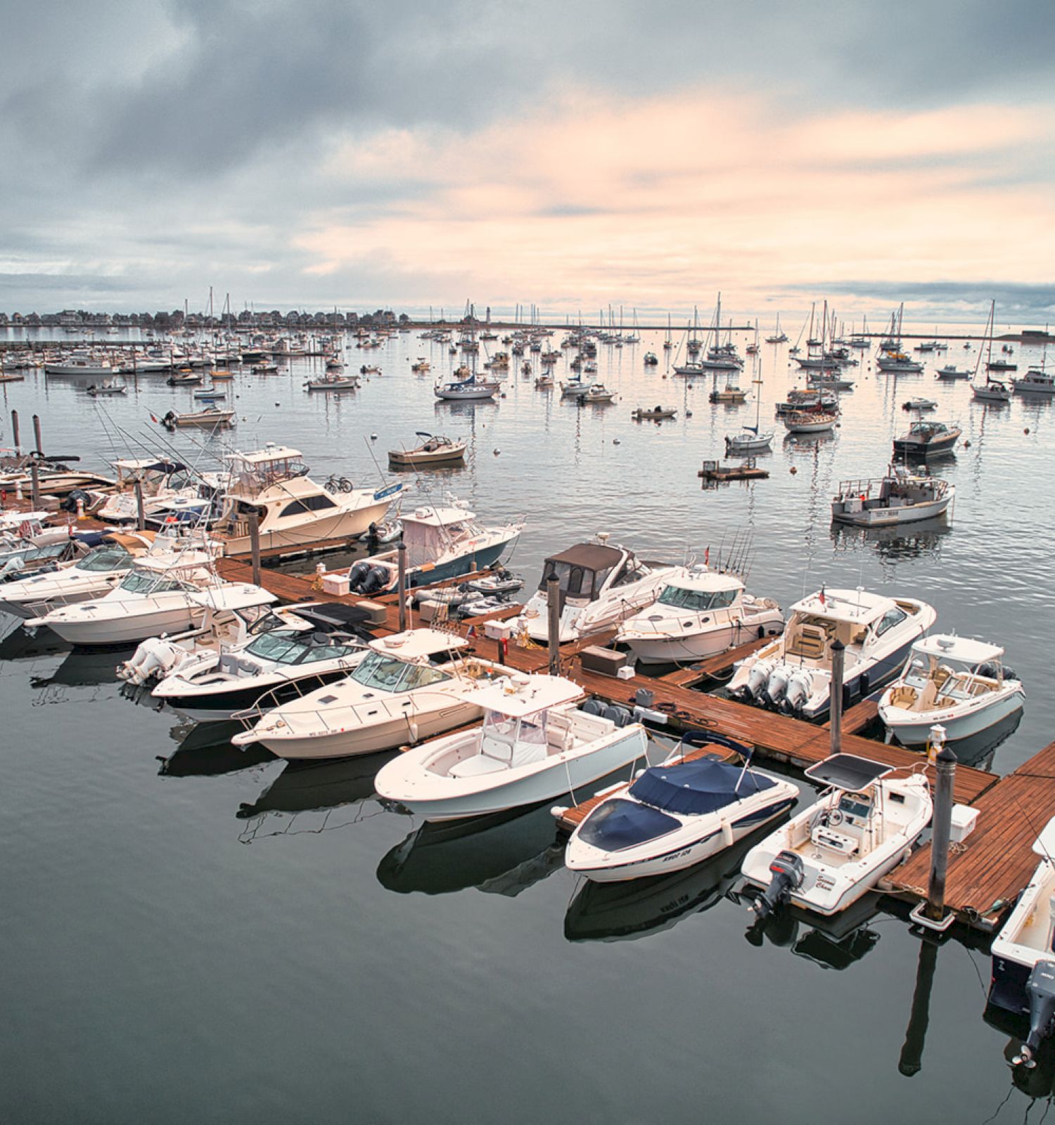 The image shows a marina with boats docked at a pier, surrounded by more boats anchored in the harbor under a cloudy sky at sunset.