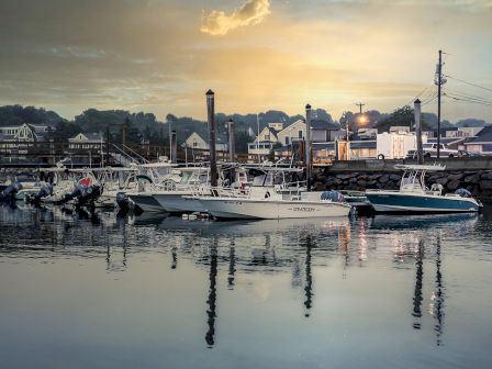 The image shows boats docked at a marina in a small coastal town during sunset. The sky is partly cloudy with soft, warm lighting reflected in the water.