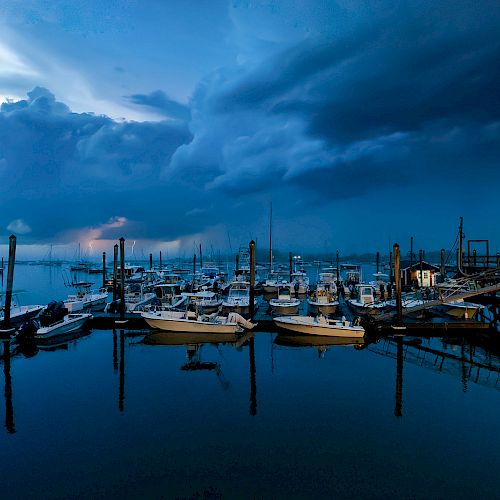 A harbor at dusk with docked boats and a dramatic sky filled with dark clouds, reflecting on the calm water, offering a serene view.